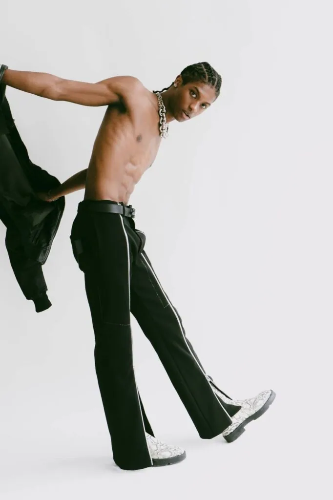 Shirtless male model in black pants and white sneakers posing dynamically against white background