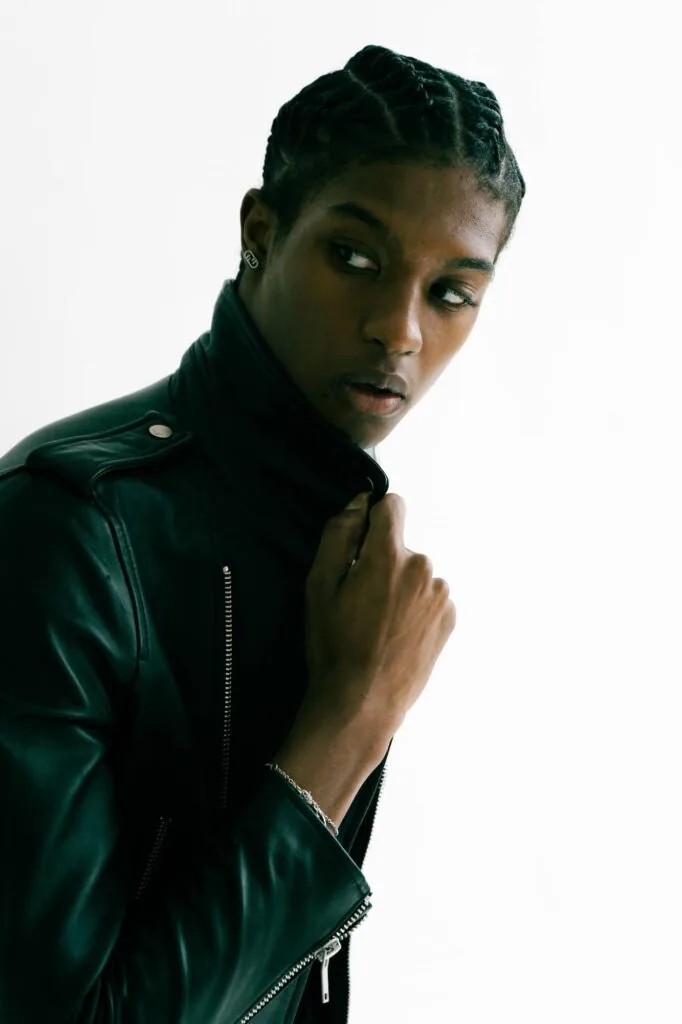 Male model wearing black leather jacket with braided hairstyle against white background