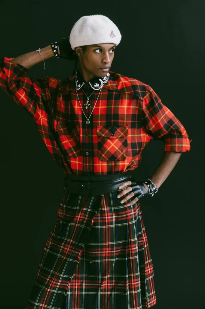 Male model wearing red and black plaid shirt, white beret, studded jewelry, and green plaid skirt against dark background.