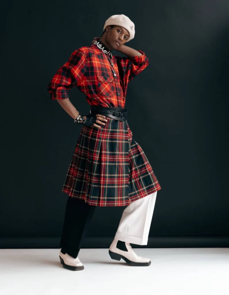 Male model in red plaid shirt, matching skirt, white beanie, and two-tone shoes posing against dark background.
