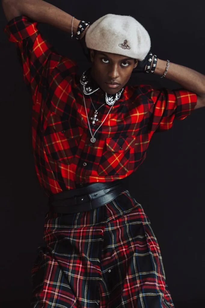 Man in red plaid shirt and matching skirt, white beret, silver jewelry against black background