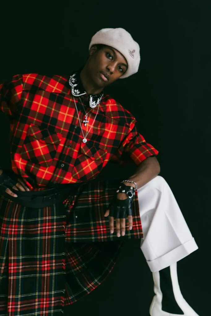 Male model in red tartan shirt, white beanie, and layered jewelry poses against dark background