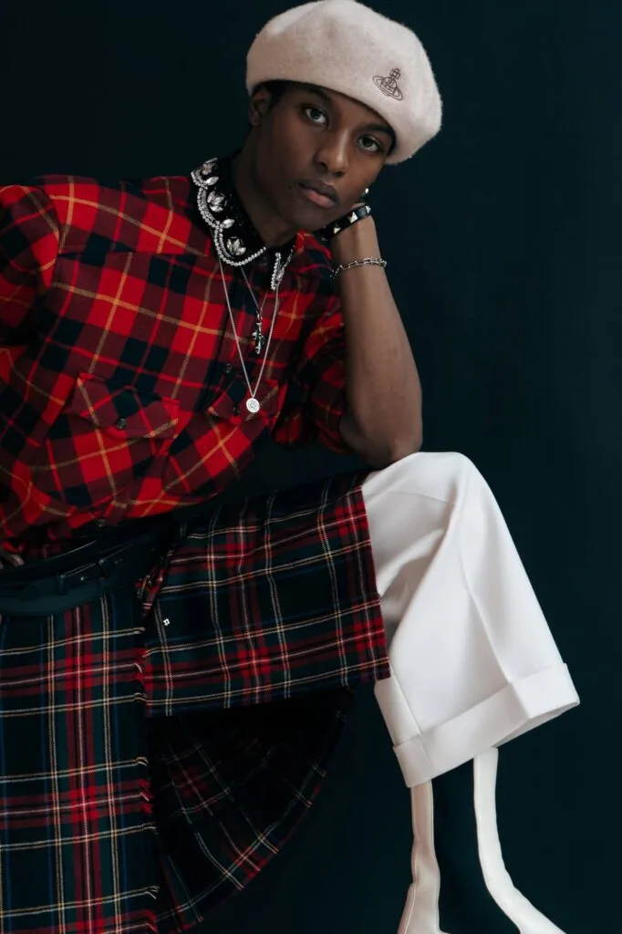 Male model wearing red plaid shirt, cream beret, silver chain necklaces, and white pants against dark background