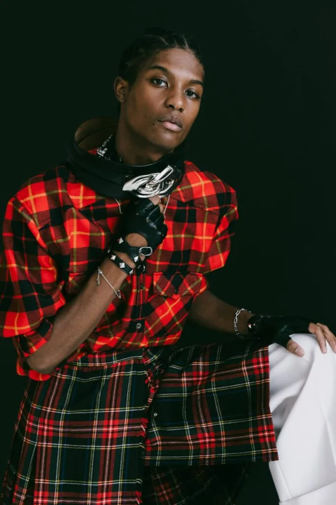 Male model wearing red and black plaid shirt with black hoodie and silver chain necklace against dark green background