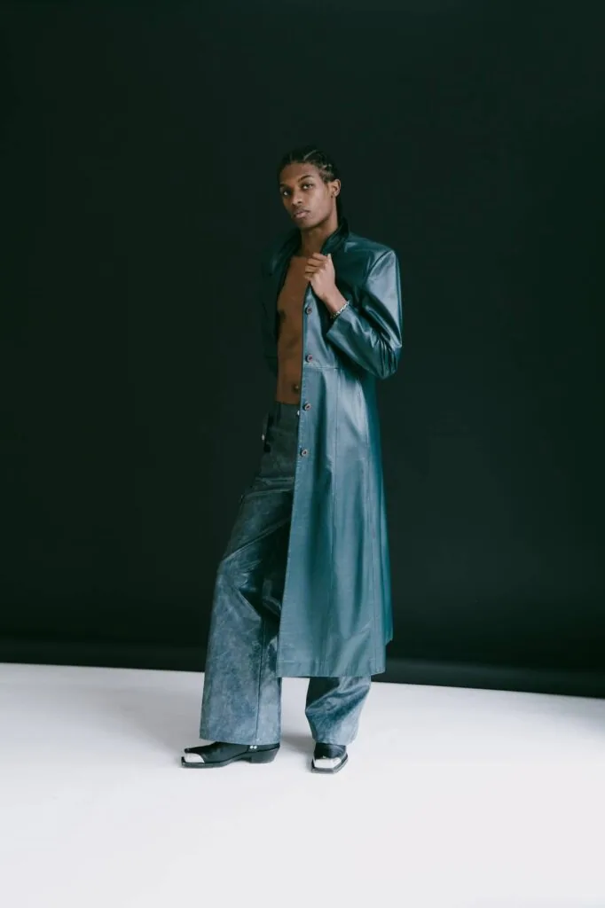 Male model wearing light blue denim coat over dark jeans and dark sneakers, posed against dark green and white studio backdrop