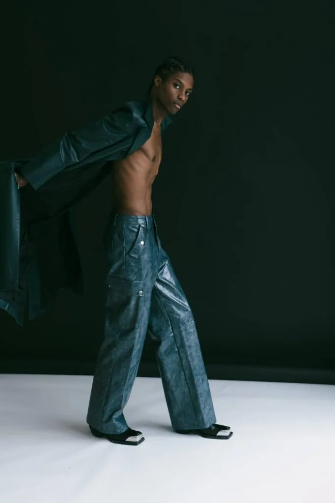 Male model shirtless wearing teal jacket and dark denim jeans against dark background