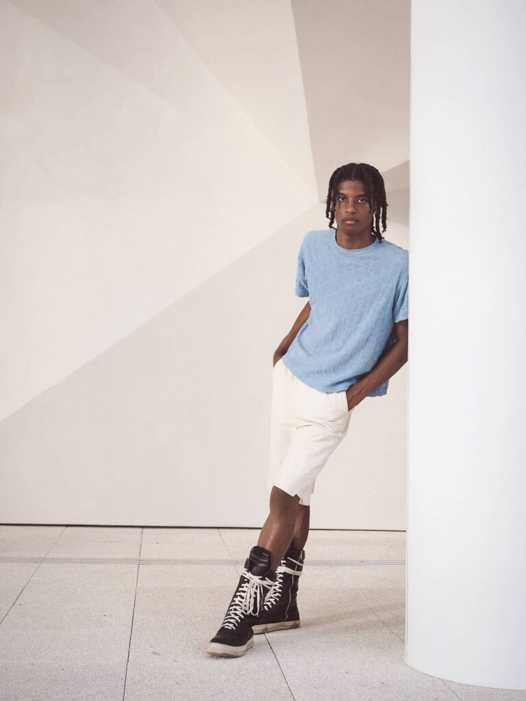 Male model wearing light blue t-shirt and white pants with black lace-up boots in minimalist gallery setting