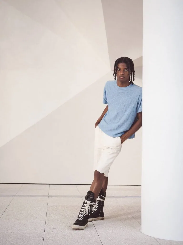 Male model wearing light blue t-shirt and white pants with black lace-up boots in minimalist gallery setting