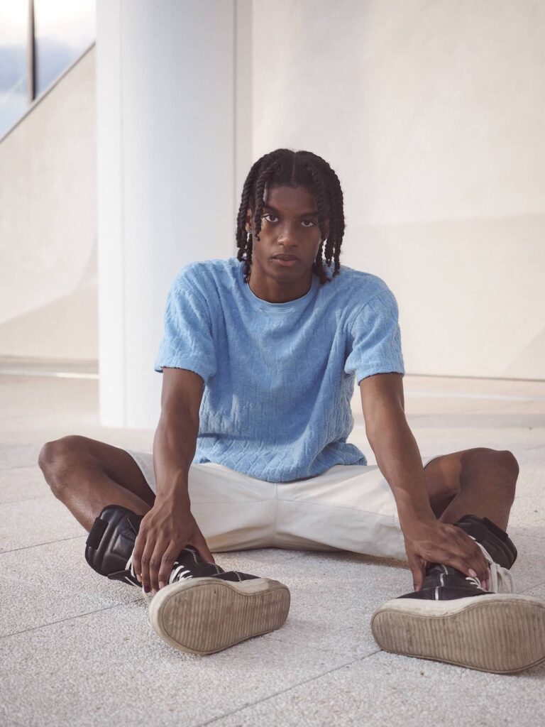 Male model in light blue t-shirt and white pants sitting cross-legged in modern white gallery space