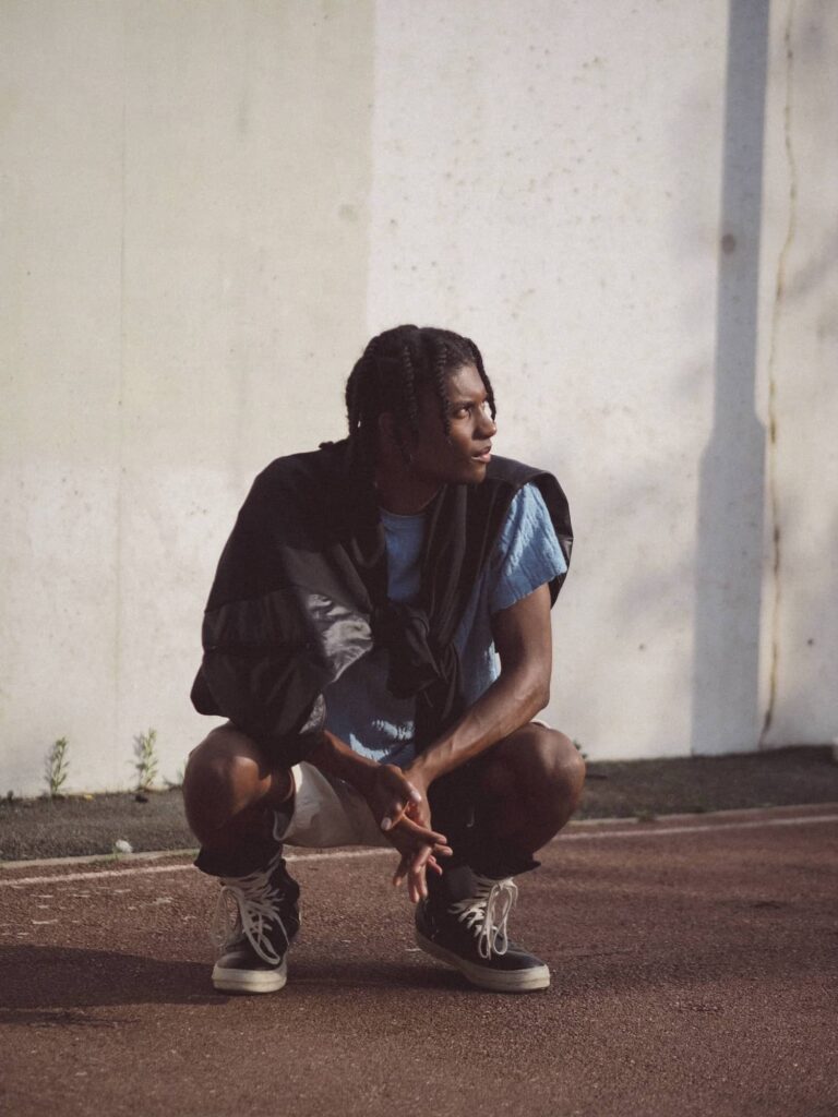 Man in black vest and light blue shirt crouching against white wall with shadow