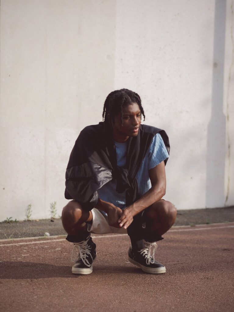 Man in black jacket and blue shirt crouching against white wall, wearing white sneakers