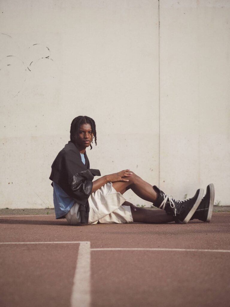 Young man with braids sitting on concrete wearing black jacket and white shorts against white wall