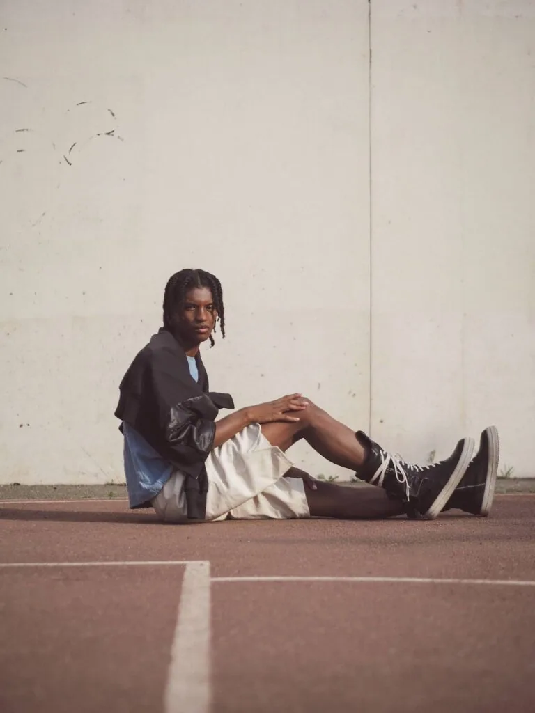 Young man with braids sitting on concrete wearing black jacket and white shorts against white wall