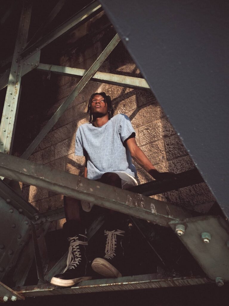 Person wearing light blue t-shirt and lace-up boots sitting on metal industrial structure with dramatic shadows