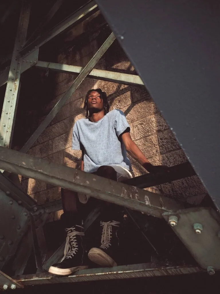 Person wearing light blue t-shirt and lace-up boots sitting on metal industrial structure with dramatic shadows