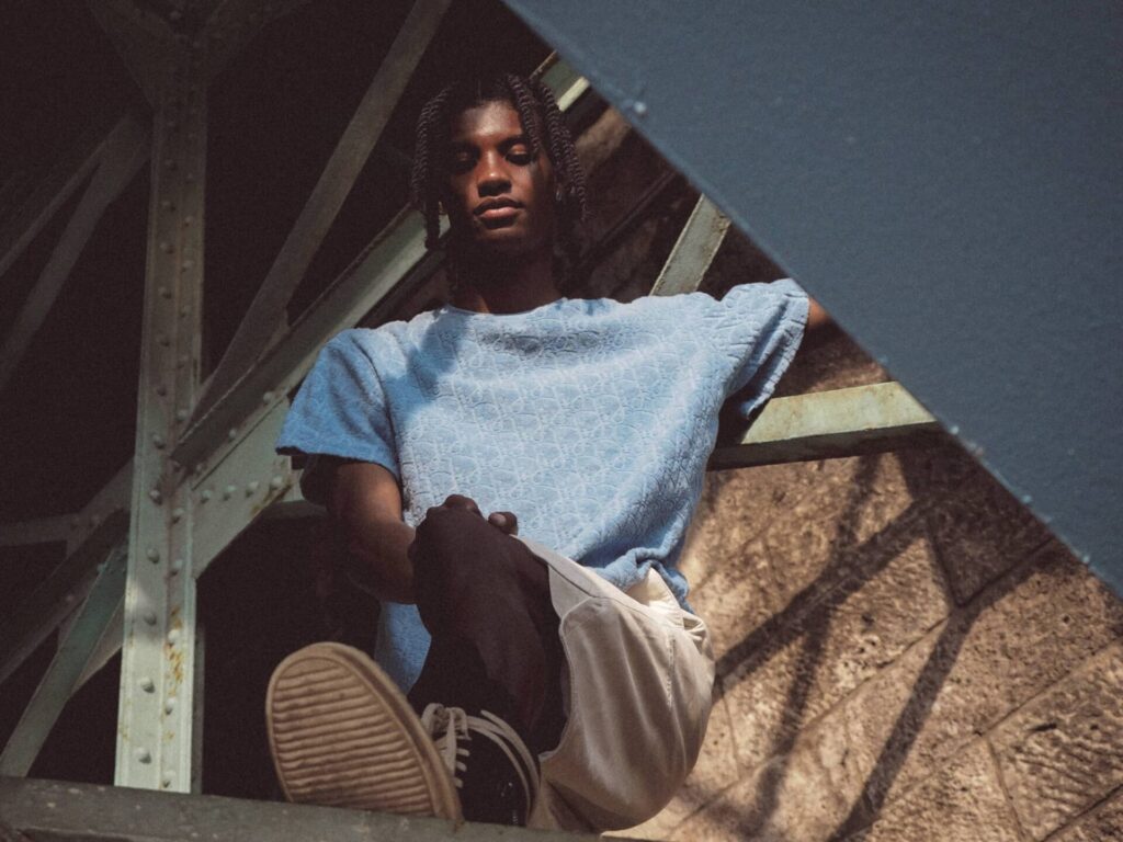 Male model in light blue t-shirt and beige pants sitting under industrial metal bridge structure with dramatic shadow