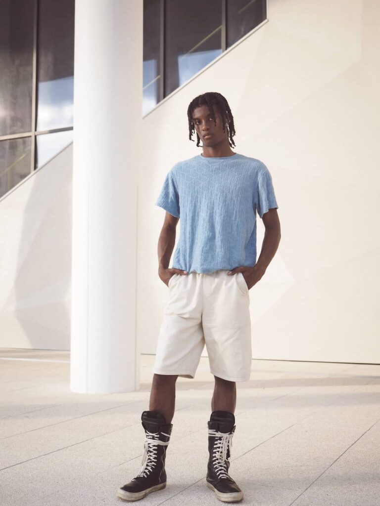 Male model wearing light blue t-shirt and white shorts with black high-top sneakers, standing in modern gallery space with white columns and large windows