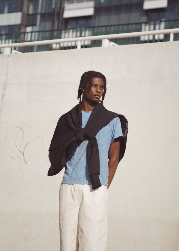 Male model wearing light blue t-shirt, black cardigan, and cream pants standing against white wall with industrial building backdrop.