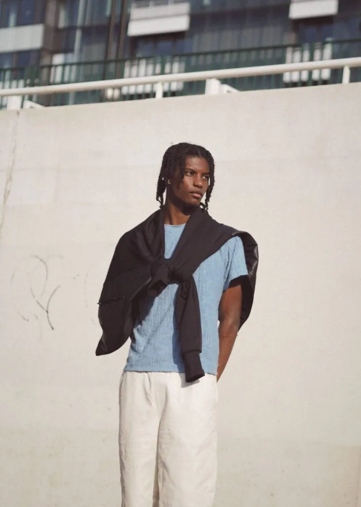 Male model wearing light blue t-shirt, black cardigan, and cream pants standing against white wall with industrial building backdrop.