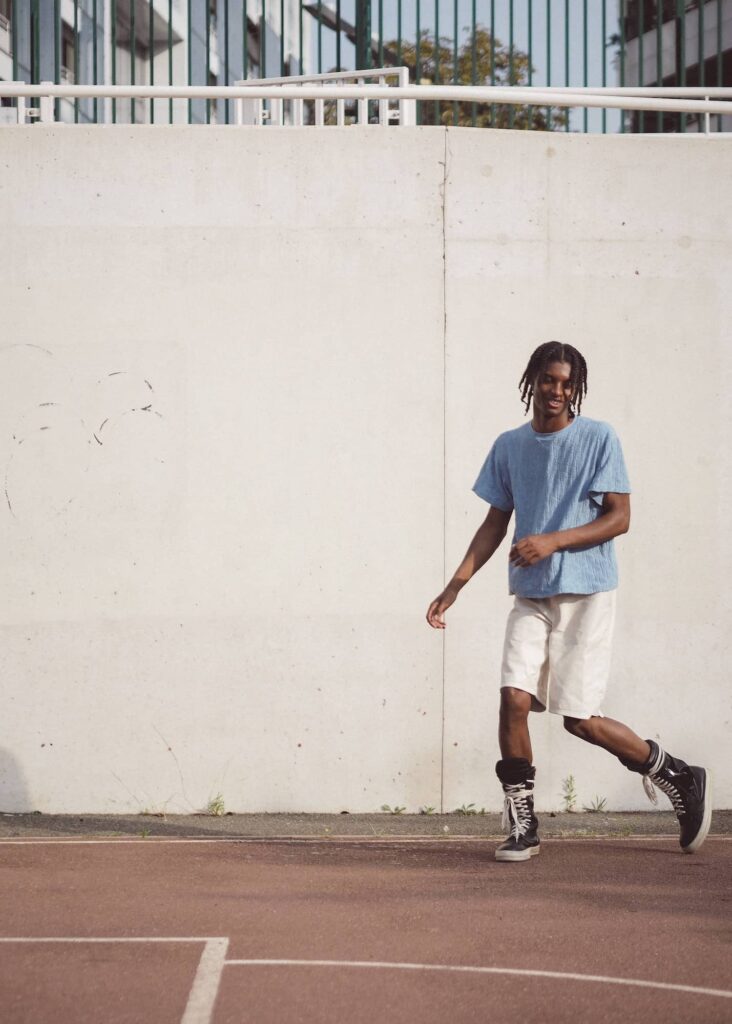 Male model in blue t-shirt and cream shorts jogging on clay court near concrete wall
