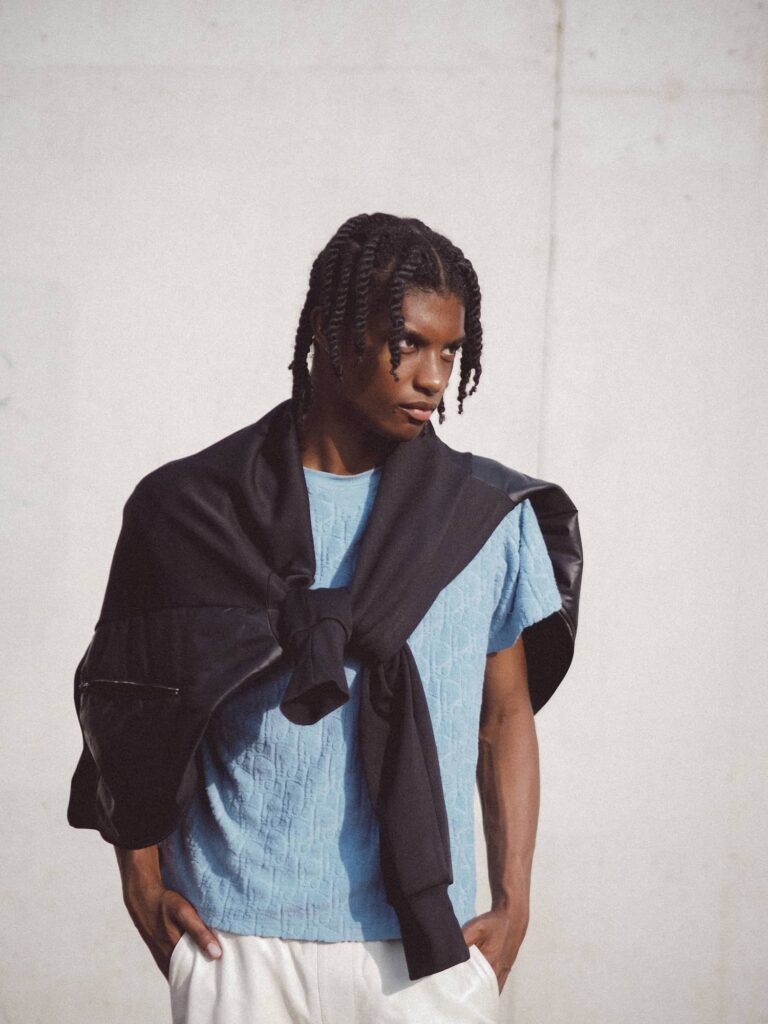 Male model wearing light blue textured t-shirt and black cardigan with white pants against white wall