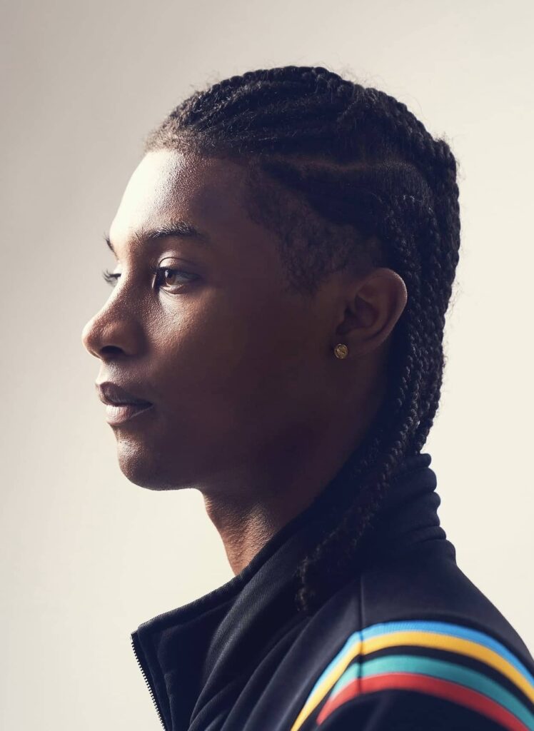 Male model in profile wearing black jacket with colorful striped sleeve detail and gold earring against neutral background