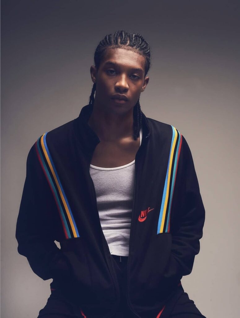 Male model wearing black track jacket with rainbow stripe trim and white undershirt, braided hairstyle, studio portrait against gray background