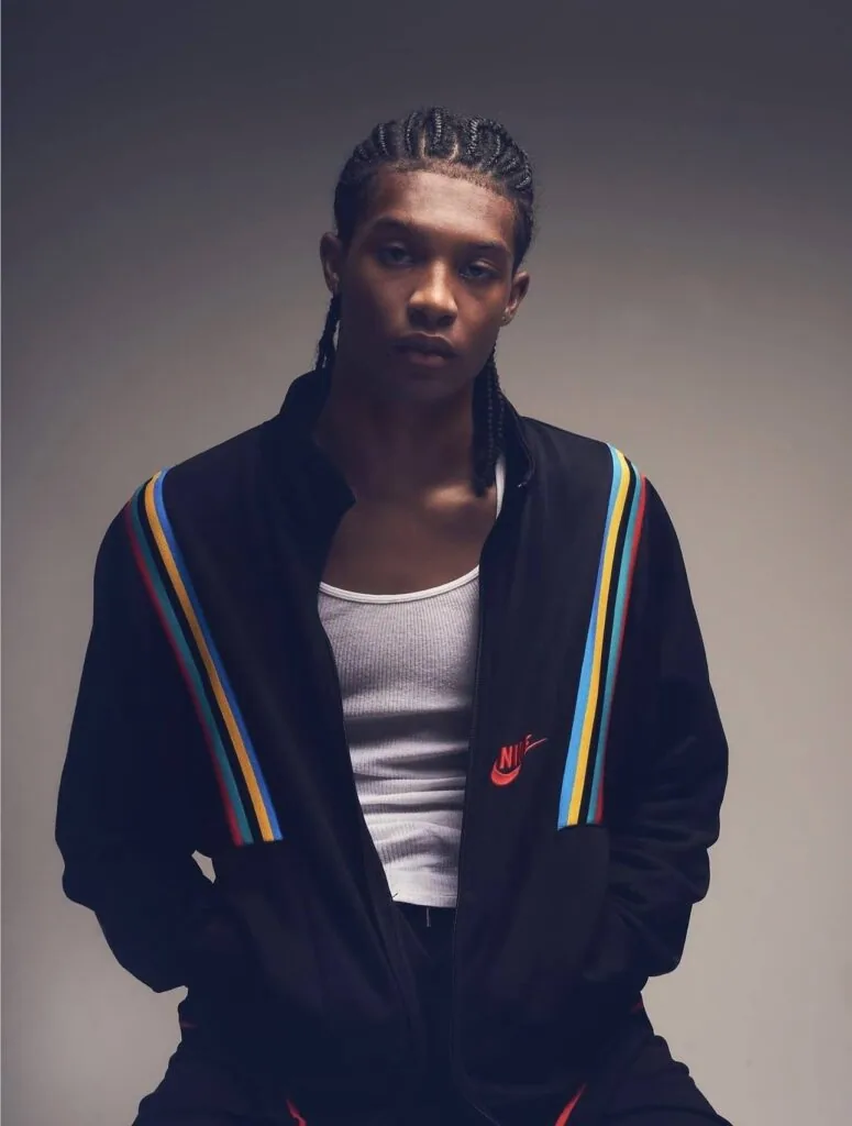 Male model wearing black track jacket with rainbow stripe trim and white undershirt, braided hairstyle, studio portrait against gray background