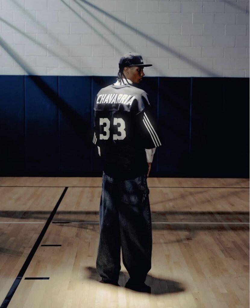 Male model wearing black Adidas jersey number 33 and dark jeans standing in gymnasium with shadow cast on wall