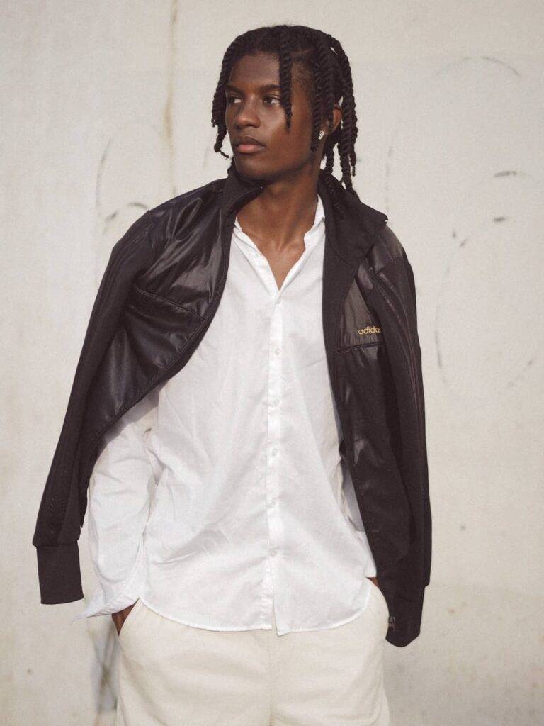 Male model wearing black jacket over white button-up shirt and cream trousers against white wall