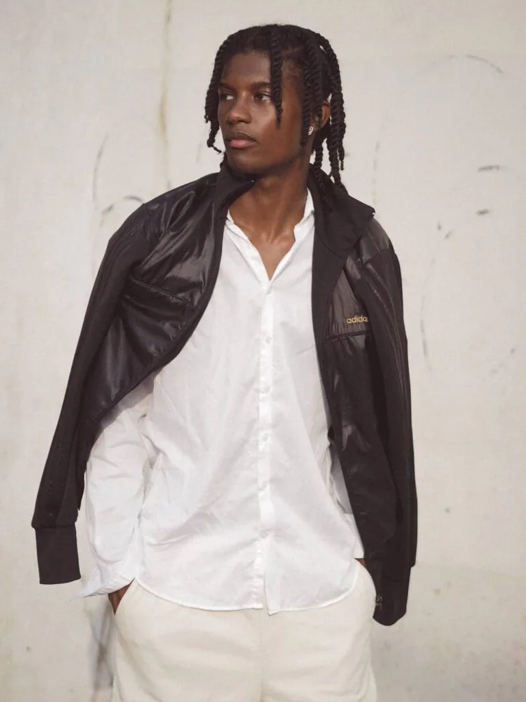Male model wearing black jacket over white button-up shirt and cream trousers against white wall