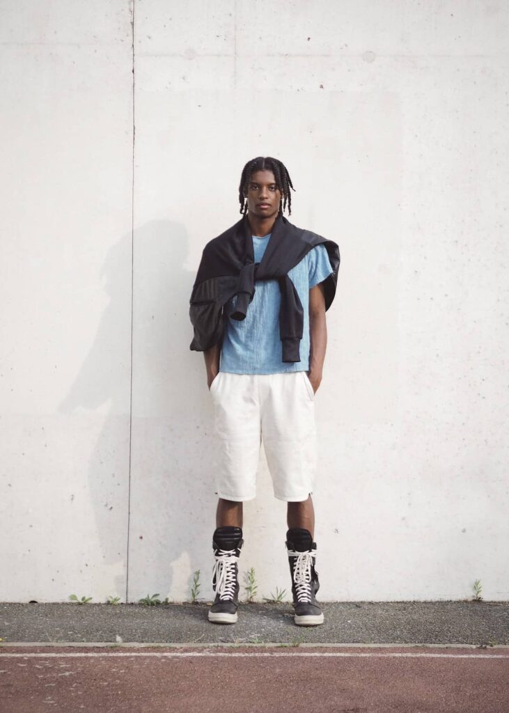Male model wearing light blue t-shirt, white shorts, black cardigan, and black high-top sneakers standing against white wall