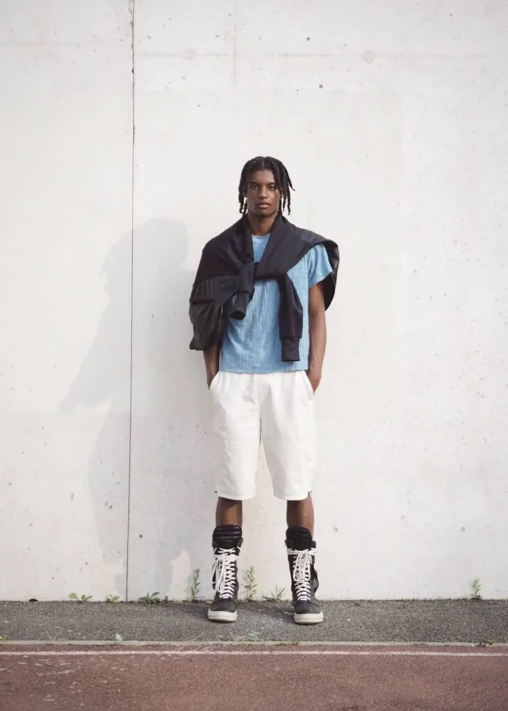 Male model wearing light blue t-shirt, white shorts, black cardigan, and black high-top sneakers standing against white wall