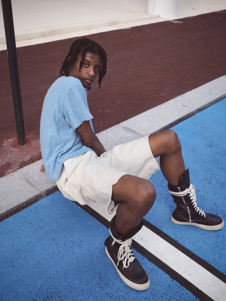 Young man in light blue t-shirt and white shorts wearing black and white high-top sneakers, sitting on blue ground with burgundy wall background.