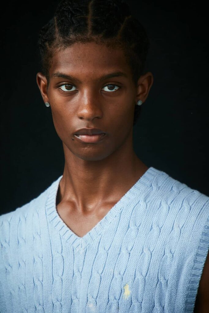 Young man with pearl earrings wearing light blue cable-knit sweater against dark background