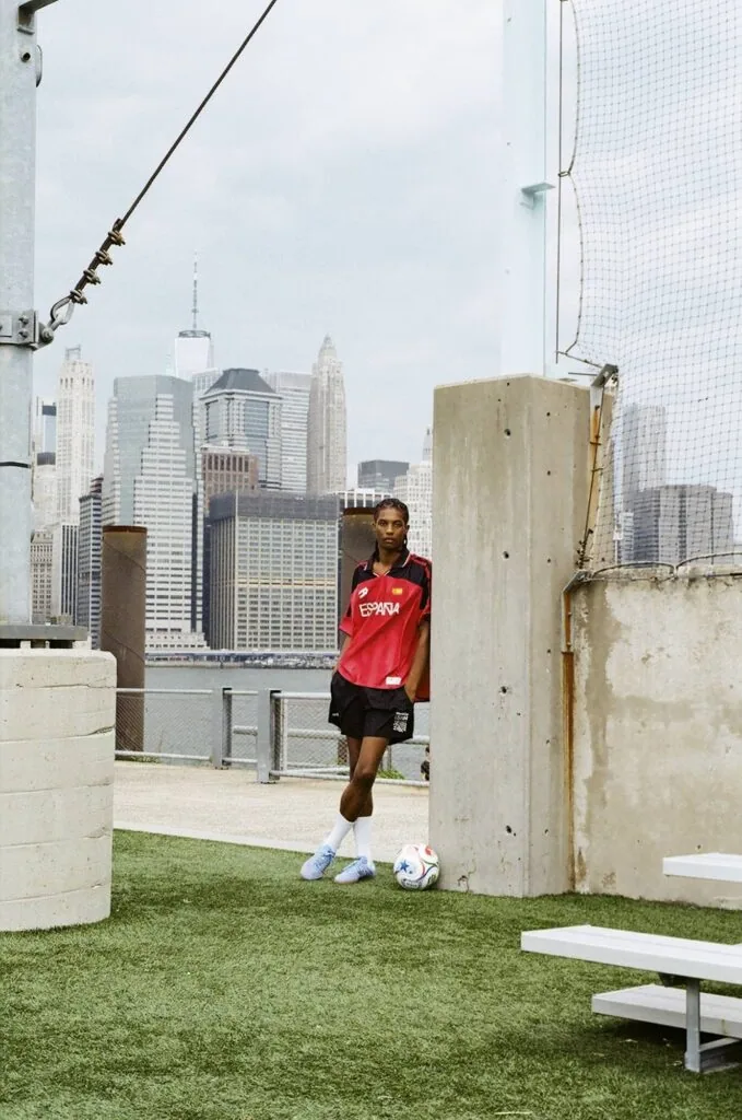 Soccer player in red jersey standing on artificial turf field with Manhattan skyline in background