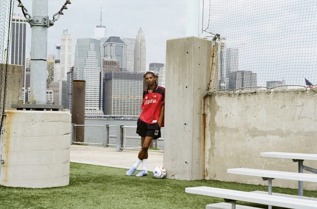 Soccer player in red shirt posing with ball on rooftop field with city skyline behind