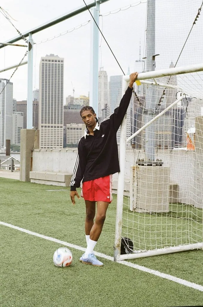 Man in black jacket and red shorts stretching on rooftop soccer field with city skyline behind him