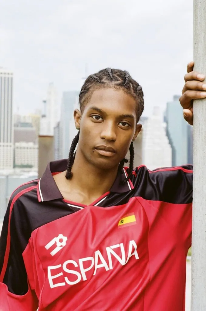 Young person wearing red and black España sports jersey leaning against white pole with city skyline behind