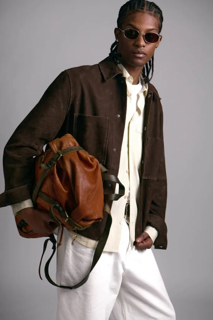 Person wearing brown suede jacket, white shirt, white pants, round sunglasses, and carrying cognac leather backpack against gray background.