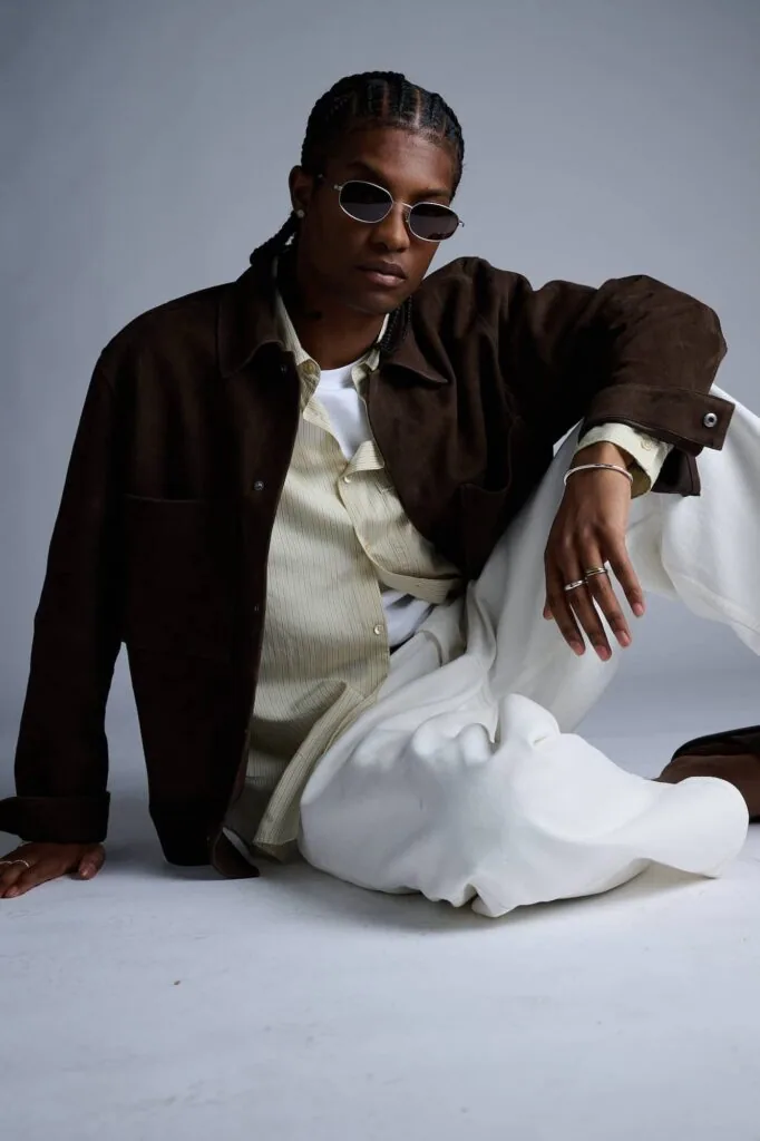 Person wearing round sunglasses, brown suede jacket, and cream shirt, posed sitting against gray background