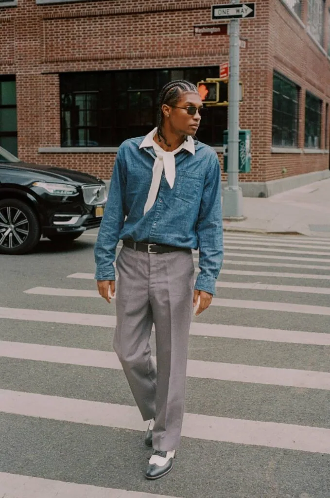 Person wearing denim shirt and gray trousers standing in crosswalk on urban street with brick buildings behind
