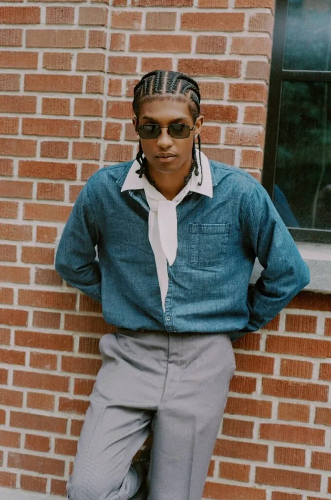 Person wearing denim shirt and gray pants leaning against red brick wall with sunglasses