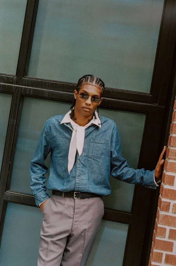 Person wearing denim shirt, white neck scarf, and light pants standing against green paneled wall with brick visible on right