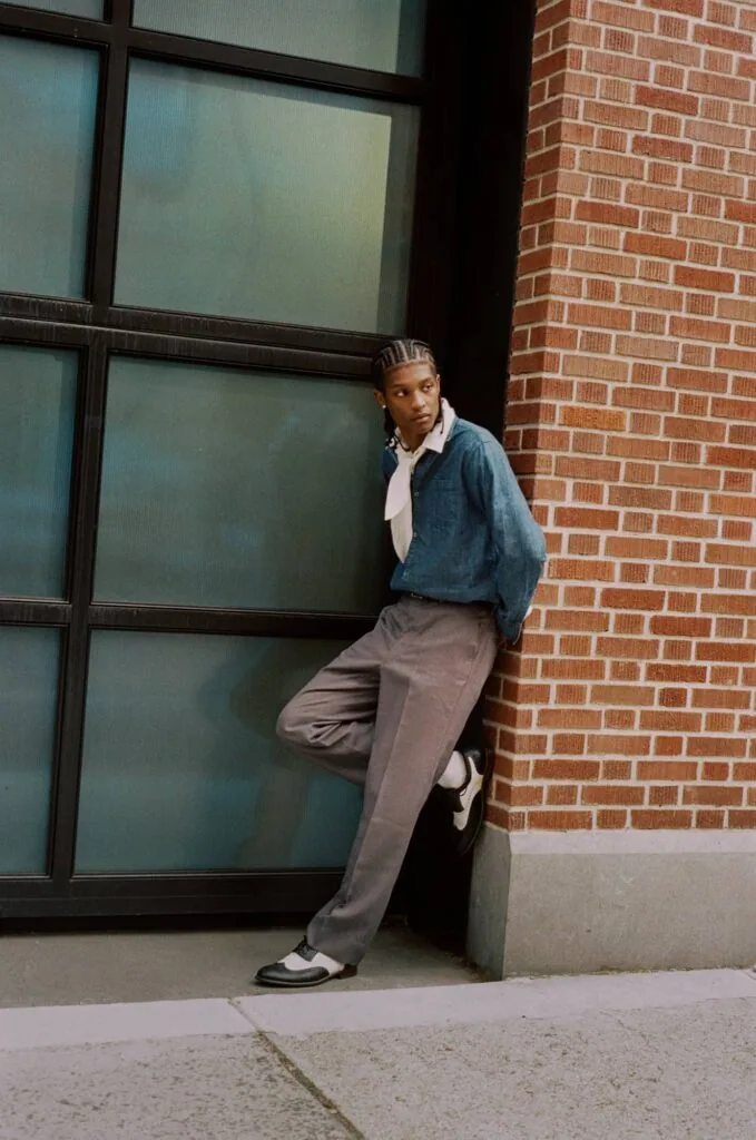 Person in denim shirt and gray pants leaning against brick wall next to large window