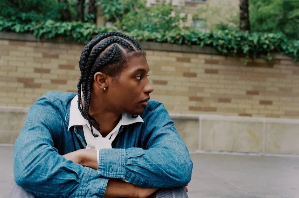 Person with braided hair wearing a denim shirt, sitting against a brick wall with ivy in the background