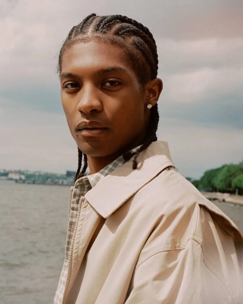 Person with braided hair wearing a beige jacket over a checkered shirt, standing by a waterfront with cloudy sky and trees in background