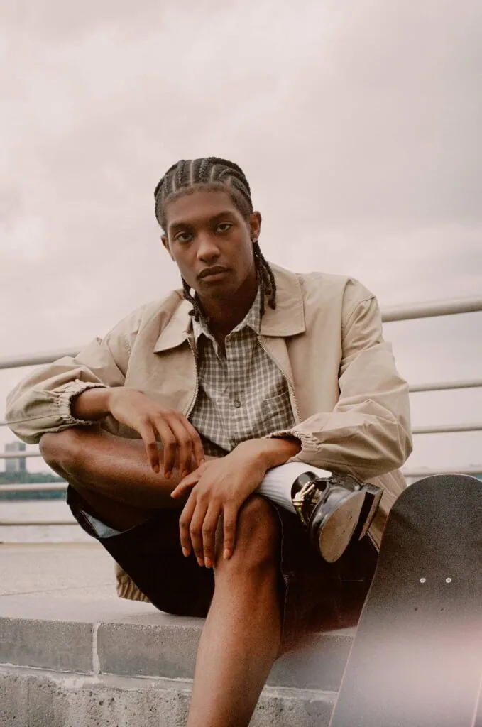 Man with braided hair wearing beige jacket and checkered shirt crouching outdoors with overcast sky behind him
