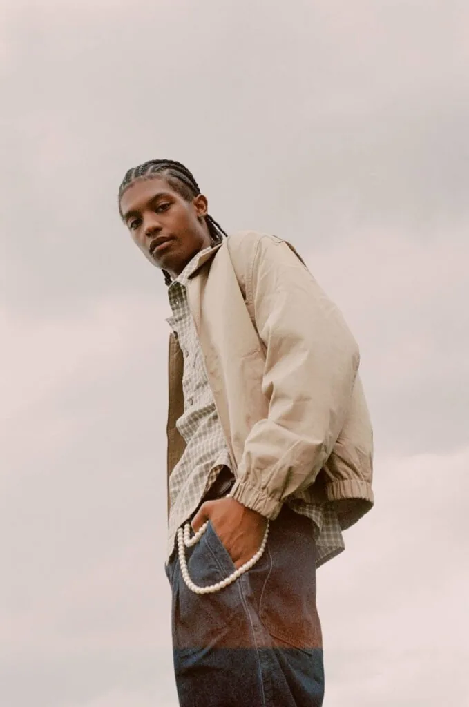 Person wearing beige windbreaker jacket over checkered shirt and dark jeans, posed against overcast sky
