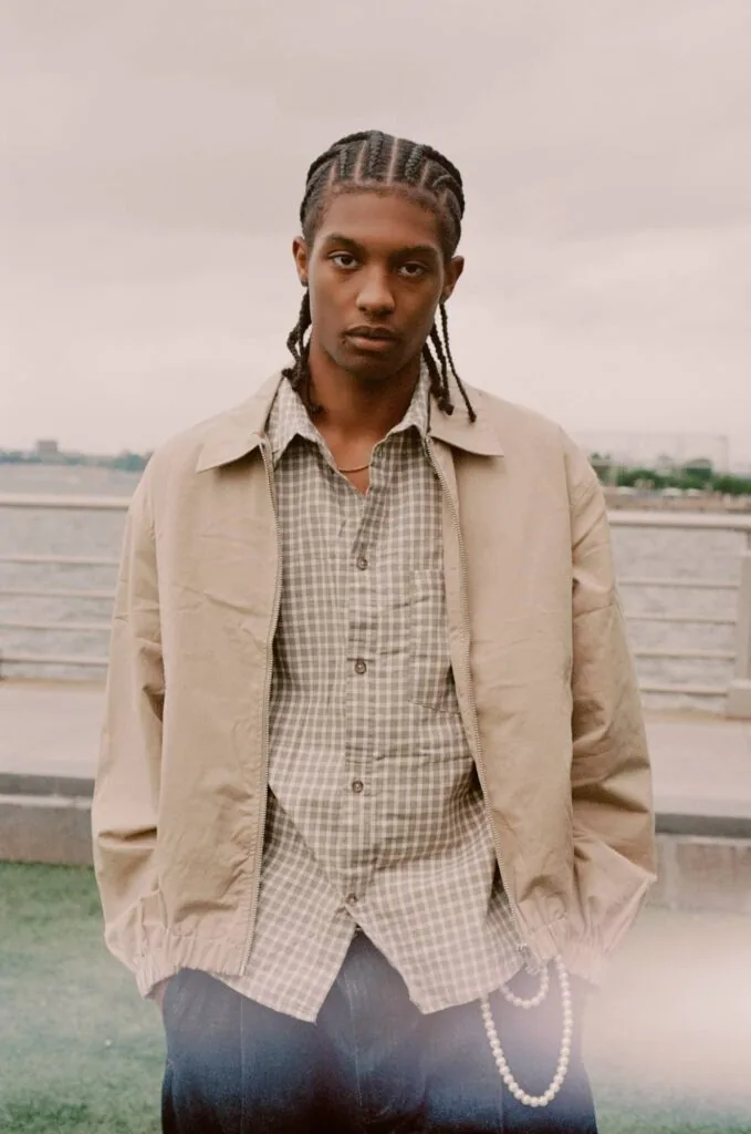 Man with braided hair wearing beige jacket and checkered shirt standing by waterfront
